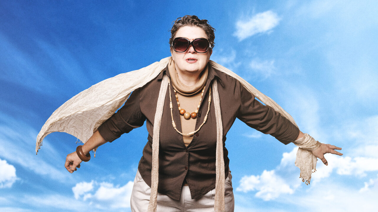 A woman in big sunglasses wearing brown and beige, with arms outstretched, her scarf billowing against a blue sky with white clouds.