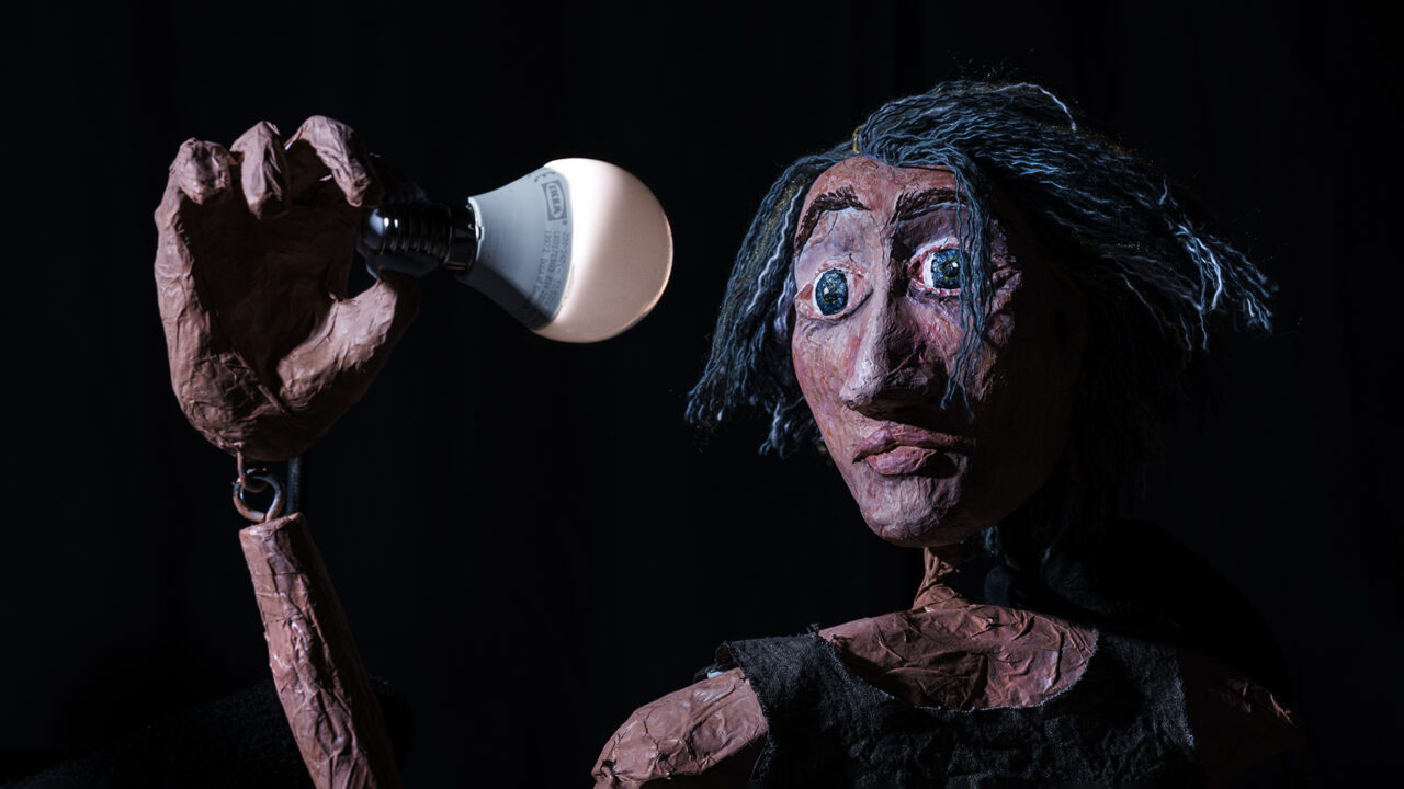 A puppet wearing a black singlet stares wide-eyed at a lightbulb