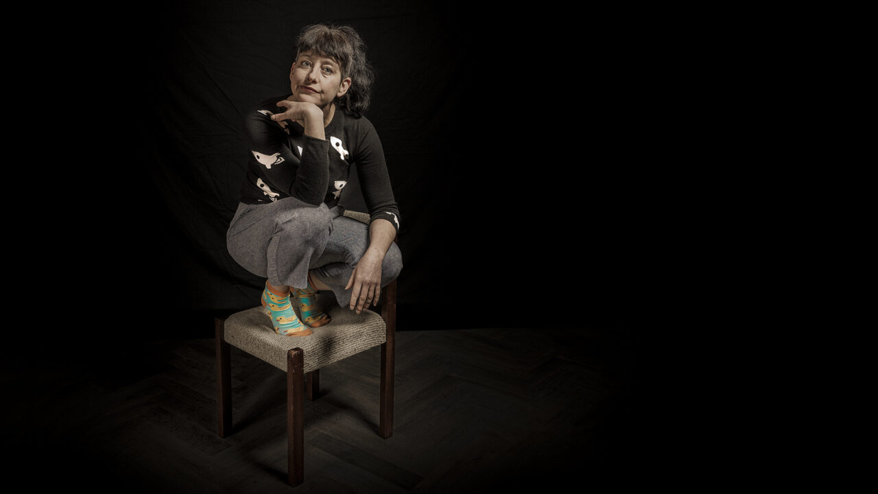 A woman, spotlit, wearing top, trousers and socks, squats on a 1980s style wooden chair. She is surrounded by complete darkness and looks bored.
