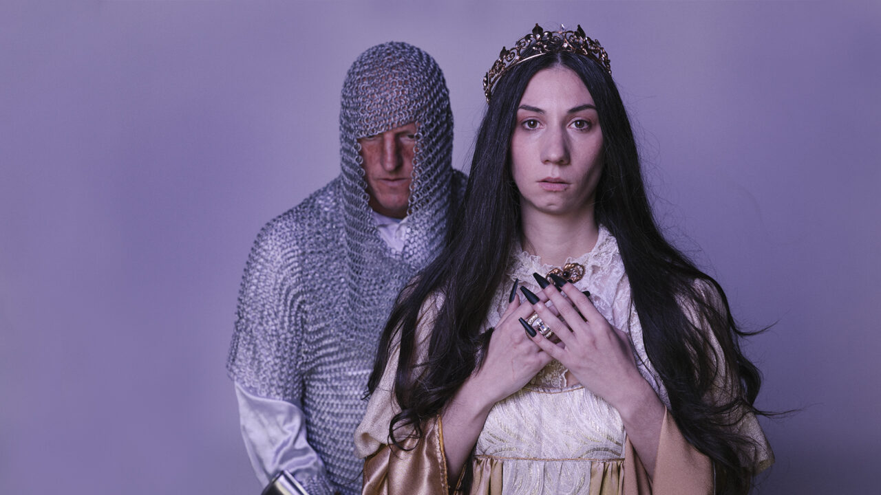 Kerith stands in the foreground in a regal dress. David in background in chainmail