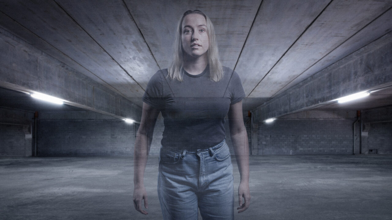 a woman with shoulder length blonde hair stands in an underground carpark. She is faded out in the image. The image is grey and industrial looking