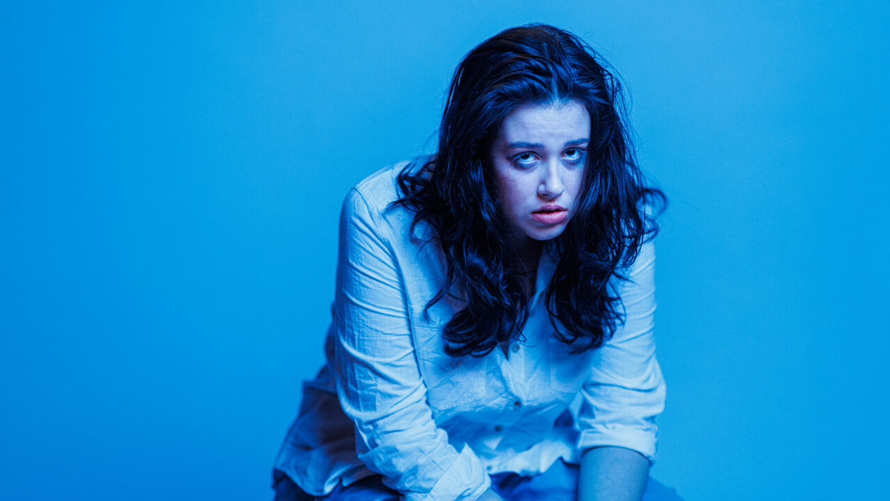 A woman with brown hair sitting on a stool staring straight at the camera. Her expression implies some pride but she has been crying. She is wearing a white button and blue jeans. Her lipstick is smudged and her eye make up is running down her face. The photo has a blue tone across the whole image.