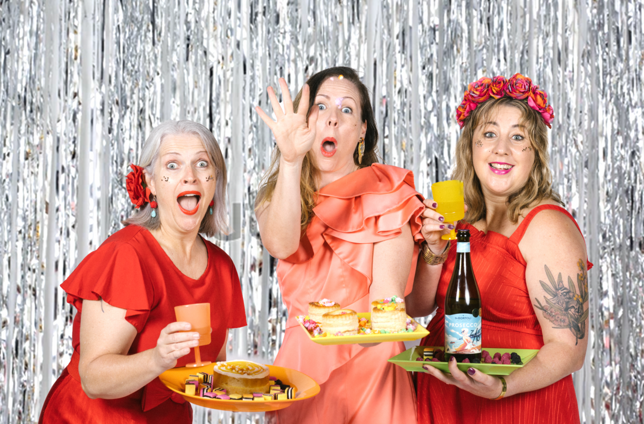 3 women stand side by side with cheeky, wide eyed, smiling expressions. They are dressed in red & salmon coloured party frocks, holding coloured platters. One Platter has a cake and licorice allsorts, one platter has vol-au-vents filled with coloured popcorn, and the last platter has a bottle of Prosecco with 4 different coloured drinking glasses. The blonde woman on the left has a purple flower pinned in her hair and red lipstick. The brown haired woman in the middle has blue eyes, red lipstick and is tossing coloured popcorn at the camera. The last woman on the right has honey blonde hair and a flower crown on her head, she is wearing purple lipstick. The background to this colourful image is silver tinsel.