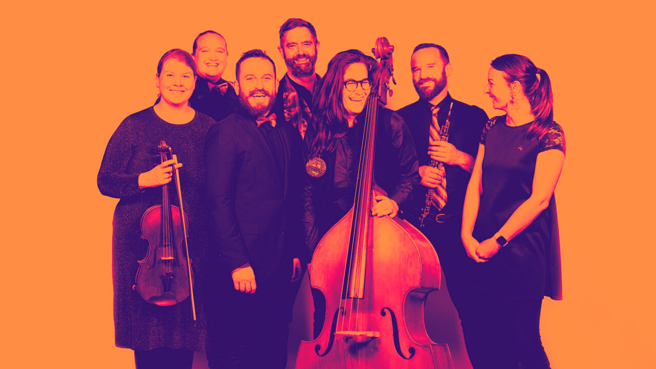 6 people gather together, smiling and laughing. Some hold musical instruments.