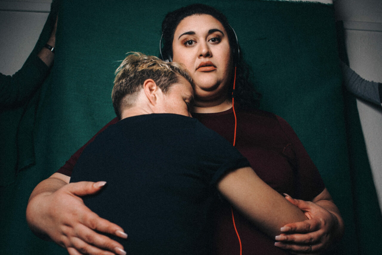 Two women. One faces away from the camera hugged by the other woman who looks into the lens.