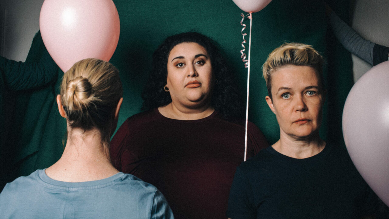 Three women stand in front of a green backdrop, two of them hold balloons, one woman has her back turned.