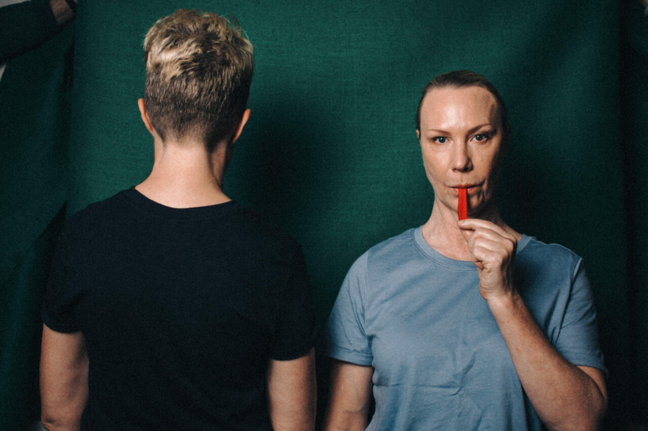 Two women stand in front of a green backdrop. One woman faces away, the second woman sucks on a red lolly.