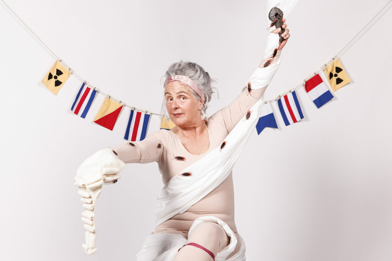 A woman in a flesh coloured unitard is wrapped in circus silks. Cockroaches crawl from a cross in her left hand, across her body to a legal wig in her right hand. She looks at us with powerful good humour.