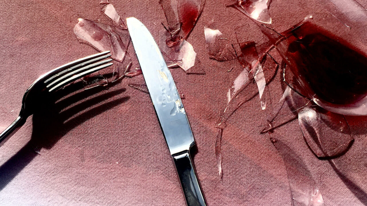 A wine glass shattered on a white table cloth with red wine seeping across the table.