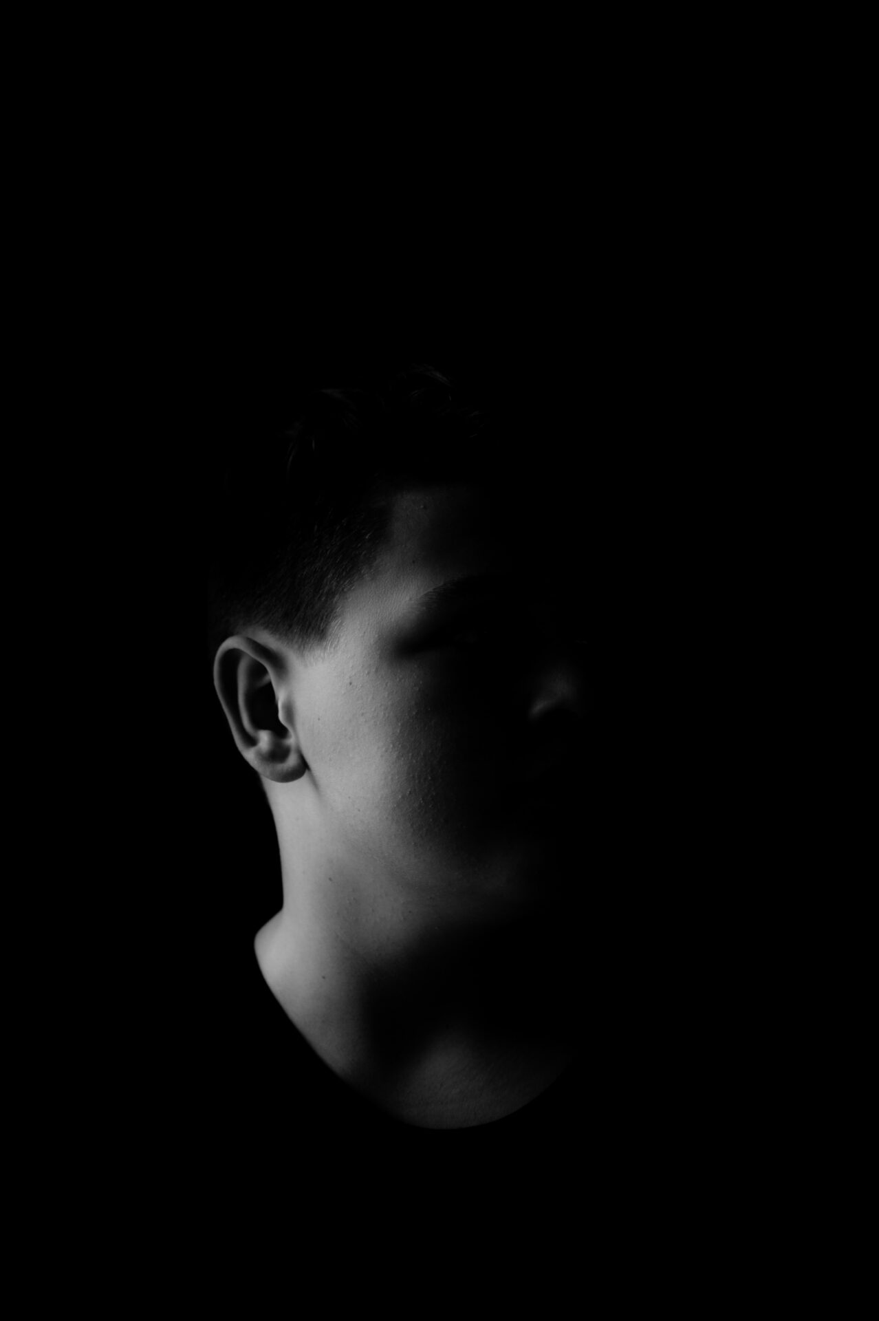 A person with short, dark hair: only one side of their face and ear is illuminated, the rest of their features disappear into shadow.